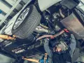 Mechanik pracuje pod samochodem, naprawiając budowę układu kierowniczego. Widać podniesione auto, koło i elementy wydechu.