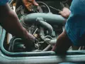 Mechanicy pracują przy silniku, jeden z nich trzyma wtyczkę, drugi dokręca śrubę. Może to być naprawa zaworu par paliwa.