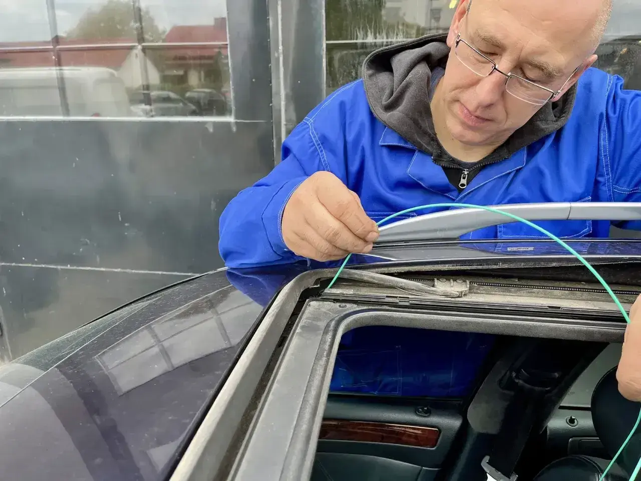 Mechanik w niebieskim kombinezonie naprawia podszybie samochodu, używając zielonego przewodu.