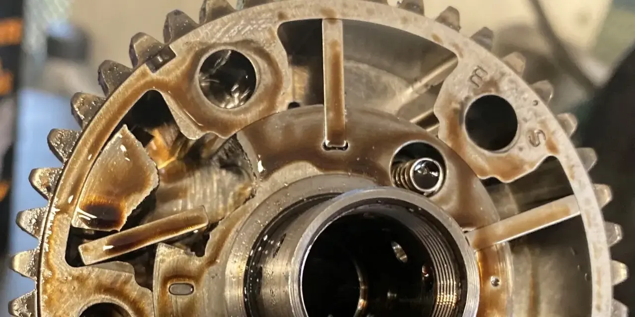 Metalowe koło zmiennych faz rozrządu, pokryte olejem, z widocznymi elementami mechanicznymi i zębatkami.