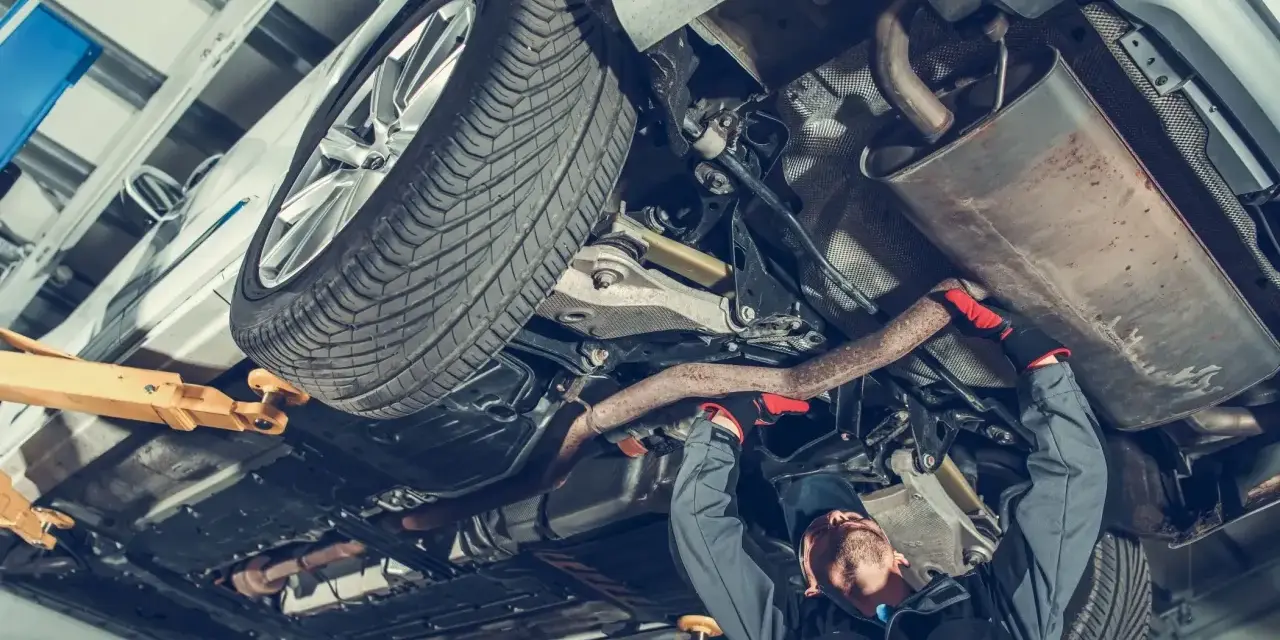 Mechanik pracuje pod samochodem, naprawiając budowę układu kierowniczego. Widać podniesione auto, koło i elementy wydechu.
