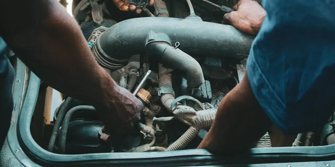 Mechanicy pracują przy silniku, jeden z nich trzyma wtyczkę, drugi dokręca śrubę. Może to być naprawa zaworu par paliwa.