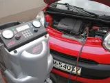 Urządzenie do obsługi klimatyzacji samochodowej podłączone do czerwonego auta. Widać manometry i klawiaturę do wprowadzania danych, a także przewody do uzupełniania czynnika chłodniczego.