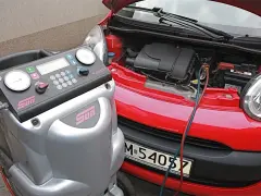 Urządzenie do obsługi klimatyzacji samochodowej podłączone do czerwonego auta. Widać manometry i klawiaturę do wprowadzania danych, a także przewody do uzupełniania czynnika chłodniczego.