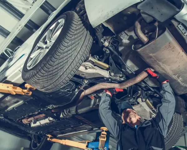 Mechanik pracuje pod samochodem, naprawiając budowę układu kierowniczego. Widać podniesione auto, koło i elementy wydechu.
