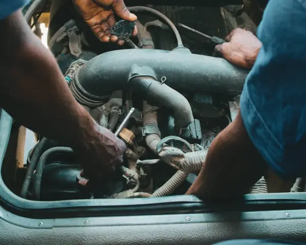 Mechanicy pracują przy silniku, jeden z nich trzyma wtyczkę, drugi dokręca śrubę. Może to być naprawa zaworu par paliwa.