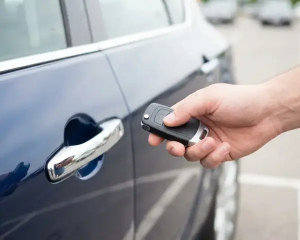 Dłoń trzyma kluczyk keyless, zbliżając go do klamki ciemnoniebieskiego samochodu.