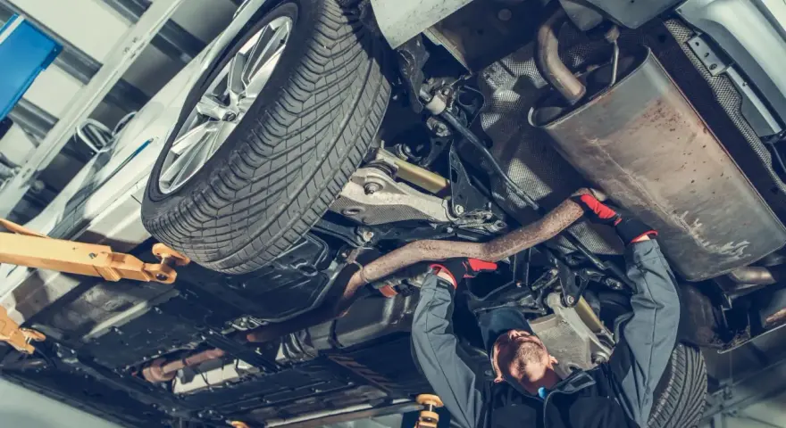 Mechanik pracuje pod samochodem, naprawiając budowę układu kierowniczego. Widać podniesione auto, koło i elementy wydechu.