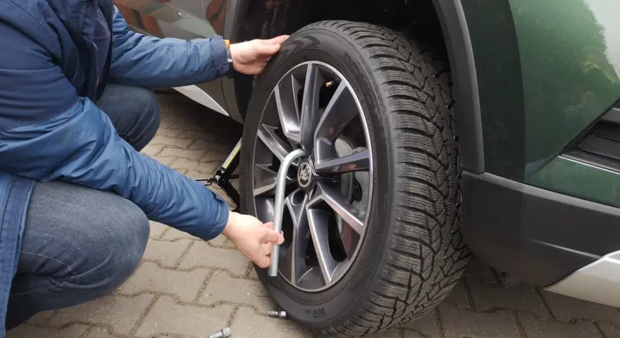 Osoba w niebieskiej kurtce dokonuje wymiany koła w samochodzie, używając klucza do odkręcenia śrub.