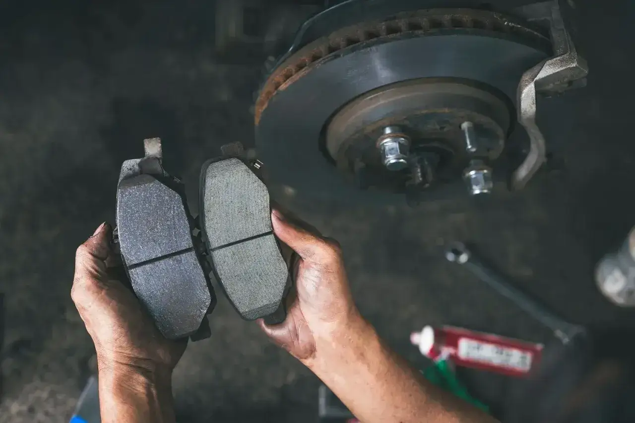 Dłonie mechanika porównują zużyte klocki hamulcowe z nowymi. W tle widać tarczę hamulcową.