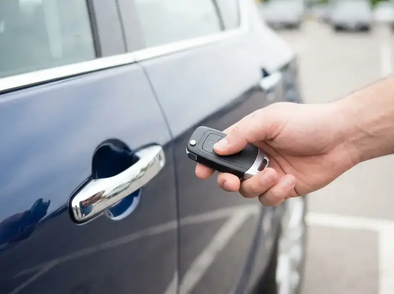 Dłoń trzyma kluczyk keyless, zbliżając go do klamki ciemnoniebieskiego samochodu.